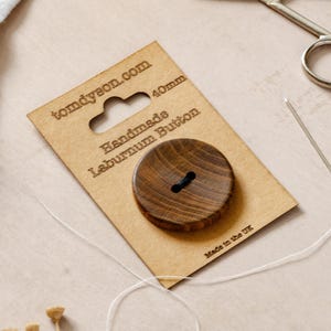 May include: A round, dark brown wooden button with a visible grain pattern, mounted on a rectangular cardboard display card labelled 'tomdyeon.com', 'Handmade Laburnum Button', and '40mm'. The card includes a needle, thread, and scissors.