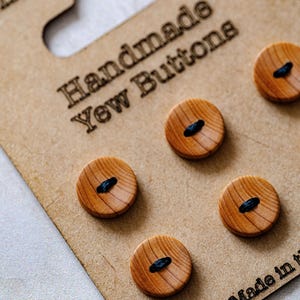 May include: A close-up shot of handmade yew wood buttons. The buttons are circular, made of light brown wood, and feature a dark blue thread detail. They are displayed on a brown cardboard backing with the text "Handmade Yew Buttons".