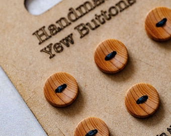 May include: Close-up of a set of handmade yew wood buttons. The buttons are round with a dark blue thread detail and are mounted on a brown cardboard backing. The text "Handmade Yew Buttons" is visible on the cardboard.