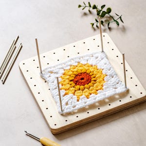 May include: A square wooden blocking board with a grid of holes and four vertical metal pins holding a square crochet motif. The motif features a central orange circle surrounded by yellow and white yarn petals. A wooden crochet hook is nearby.