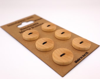 May include: A set of six light-coloured wooden buttons with black thread detail, presented on a brown cardboard backing. The buttons are evenly spaced. The cardboard has a hang hole and text that reads "Handmade Maria Buttons" and "Made in the UK".