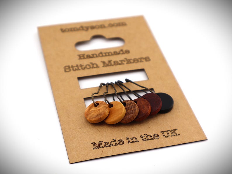 May include: Handmade stitch markers on a brown cardboard backing. The markers are made of wood in various shades of brown and black. The cardboard has the text "tomdyson.com", "Handmade Stitch Markers", and "Made in the UK".