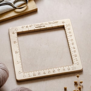 May include: A square wooden 10 cm x 10 cm Tension Ruler with etched markings and the text "4" x 4" Tension Ruler". The ruler has measurements from 0 to 10 on each side. The background includes yarn, scissors, and dried flowers.