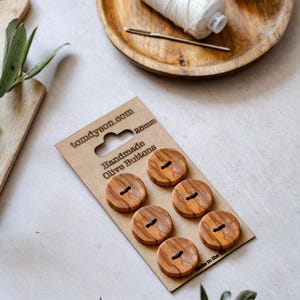 May include: A pack of six round, light brown olive wood buttons with a visible grain pattern, each featuring two holes. The buttons are mounted on a rectangular cardboard display card labelled with the text tomdyson.com, 28mm, and Handmade Olive Buttons.