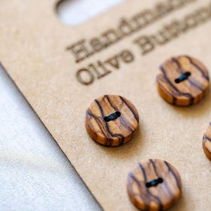 May include: Round wooden buttons with dark brown grain patterns and two centre holes, attached to a tan cardboard display card featuring the embossed text "Handmade Olive Buttons". The buttons exhibit a natural light brown wood finish.