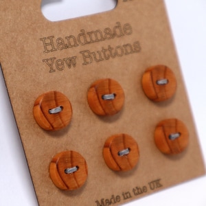 May include: A card of six handmade yew wood buttons. The buttons are circular, with a light brown hue and a visible wood grain. Each button has two holes with grey thread. The card reads "Handmade Yew Buttons" and "Made in the UK".