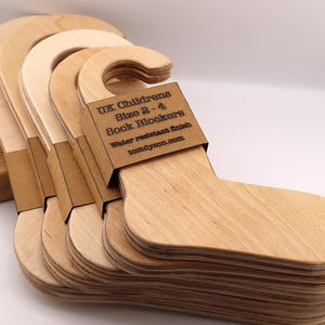 May include: A stack of light-coloured wooden sock blockers, shaped like feet, with a curved top for hanging. A brown paper label is attached to the top of the blockers, with the text "UK Childrens Size 2-4 Sock Blockers Water resistant finish tomdyson.com."