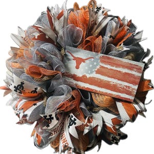 May include: A wreath decorated in orange, white, and gray mesh with a distressed American flag featuring a Longhorn steer silhouette. The wreath also includes ribbons with the words "Hook 'em Horns".