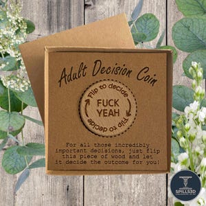 以下が含まれることがあります： 茶色の箱に入った木製の大人用意思決定コイン。「FUCK YEAH」と「Flip to decide」と書かれたコイン。箱には「非常に重要な決定は、この木の切れ端をひっくり返して結果を決定させてください！」と書かれています。