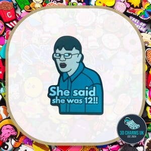 May include: A blue shoe charm featuring a cartoon character with dark hair and glasses, wearing a collared shirt. The text "She said she was 12!!" is printed in light blue letters across the lower portion of the character's torso and shirt.