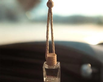 May include: A small, clear, square-shaped glass bottle with a cork stopper and a beige string for hanging. The bottle contains a clear liquid, likely a fragrance or essential oil, and is designed to be hung in a car or small space.