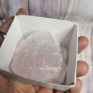 Peut inclure: Un gros cristal de quartz rose brut de couleur rose pâle dans une boîte blanche. Le cristal a une apparence légèrement nuageuse.