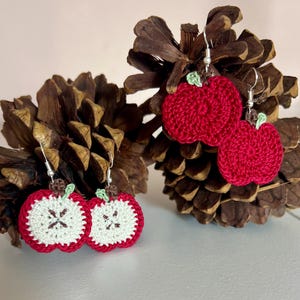 May include: Two pairs of handmade crochet apple earrings. One pair features solid red apples with small green leaves. The second pair depicts apple slices with white centers, red borders, brown stems, and green leaves, all attached to silver-toned hooks.