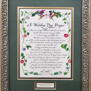 Può includere: Una preghiera di matrimonio incorniciata con un design floreale e calligrafia. Il testo recita "A Wedding Day Prayer" e include una poesia sull'amore e il matrimonio. La cornice è dorata con un passepartout verde. La parte inferiore della cornice ha una piccola targa con i nomi "Jason & Maria Spencer" e la data "December 14, 2024".