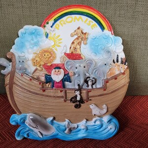 May include: A colourful ceramic wall plaque depicting Noah's Ark with a rainbow above it and the word "PROMISE" written on the rainbow. The ark is filled with animals, including lions, elephants, giraffes, and a monkey. The ark is floating on blue water with a dolphin and other sea creatures.