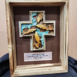 May include: A framed wooden cross with a blue background and four gold fish sculpted into the cross. The frame has a brown mount and a white inscription that reads "AND HE SAID TO THEM "FOLLOW ME, AND I WILL MAKE YOU FISHERS OF MEN" Matthew 4:19