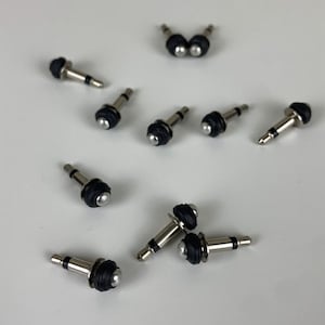 Puede incluir: Primer plano de diez conectores de audio de color plateado con puntas de goma negra. Los conectores están dispuestos de forma dispersa sobre una superficie blanca.