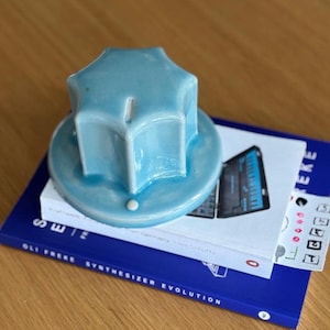 May include: A light blue ceramic knob-shaped object with a star-like top, resting on a stack of books. The books are titled "SWE" and "Oli Freke Synthesizer Evolution". The knob has a small white dot on its base.