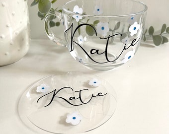Könnte beinhalten: Eine klare Glas-Tasse mit einem weißen Blumenmuster und dem Namen "Katie" in schwarzer Schrift. Die Tasse wird mit einem passenden, klaren Glasuntersetzer mit dem gleichen Design geliefert.