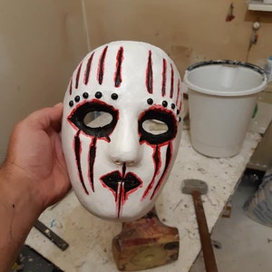 May include: White mask with black and red paint. The mask has black eyes, a black mouth, and black dots around the eyes. The mask is held in a hand.