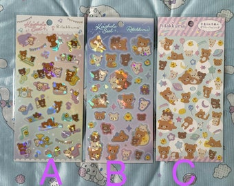 May include: Three sheets of colorful holographic stickers featuring Rilakkuma and Kiiroitori characters. The stickers are on a white background with a rainbow gradient. The stickers are labeled "Kiraholo Seal" and "Rilakkuma".