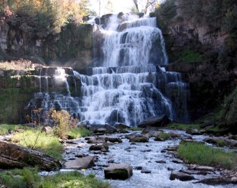 Könnte beinhalten: Ein mehrstufiger Wasserfall stürzt eine felsige Klippe hinunter in einen Bach. Das Wasser ist weiß und schäumend, im Kontrast zu den dunklen Felsen und der grünen Vegetation. Bäume mit Herbstfarben rahmen die Szene ein.
