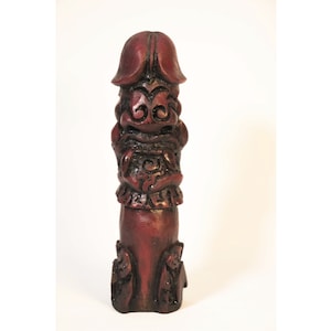May include: A carved wooden sculpture in a reddish-brown hue, featuring a stylized face and body. The sculpture has a phallic shape with detailed facial features and decorative elements. The piece is set against a plain white background.