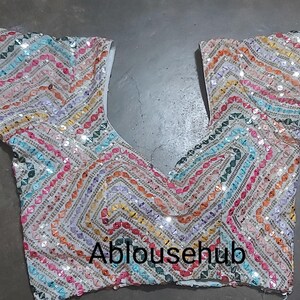 Puede incluir: Una blusa blanca con lentejuelas con un patr&oacute;n geom&eacute;trico colorido. La blusa tiene cuello en V y mangas cortas. El texto "Ablousehub" es visible en la parte delantera de la blusa.