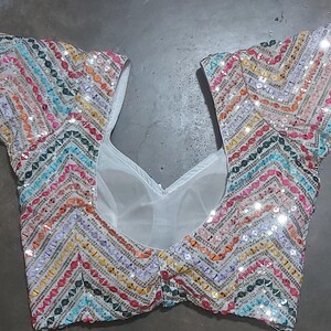 Puede incluir: Una blusa blanca con lentejuelas con un patr&oacute;n de chevron en colores arco&iacute;ris. La blusa tiene mangas cortas y un escote en V.