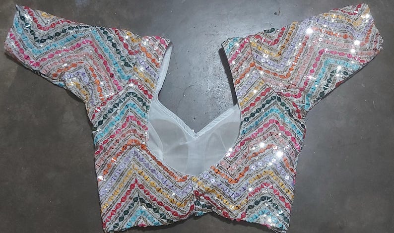 Puede incluir: Una blusa blanca con lentejuelas con un patr&oacute;n de chevron en colores arco&iacute;ris. La blusa tiene mangas cortas y un escote en V.