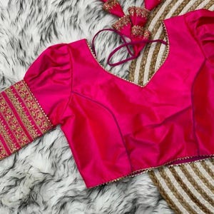 Puede incluir: Una blusa rosa fucsia sin mangas con escote en V y mangas abullonadas. Las mangas y el escote están adornados con bordados dorados.