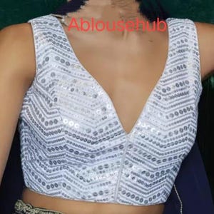 Puede incluir: Blusa blanca sin mangas con un escote en V profundo y un patr&oacute;n de lentejuelas plateadas. La blusa tiene una cremallera en la parte delantera.