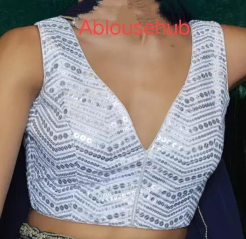 Puede incluir: Blusa blanca sin mangas con un escote en V profundo y un patr&oacute;n de lentejuelas plateadas. La blusa tiene una cremallera en la parte delantera.