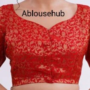 May include: A red brocade blouse with a gold floral pattern and a button closure. The blouse has a V-neckline and short sleeves. Ablousehub is written in white text on the front of the blouse.