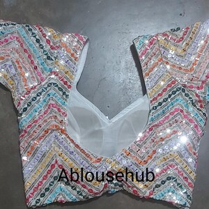 Puede incluir: Una blusa blanca con lentejuelas con un patr&oacute;n de chevron colorido. La blusa tiene un escote redondo y mangas cortas. El texto "Ablousehub" est&aacute; impreso en la parte delantera de la blusa.