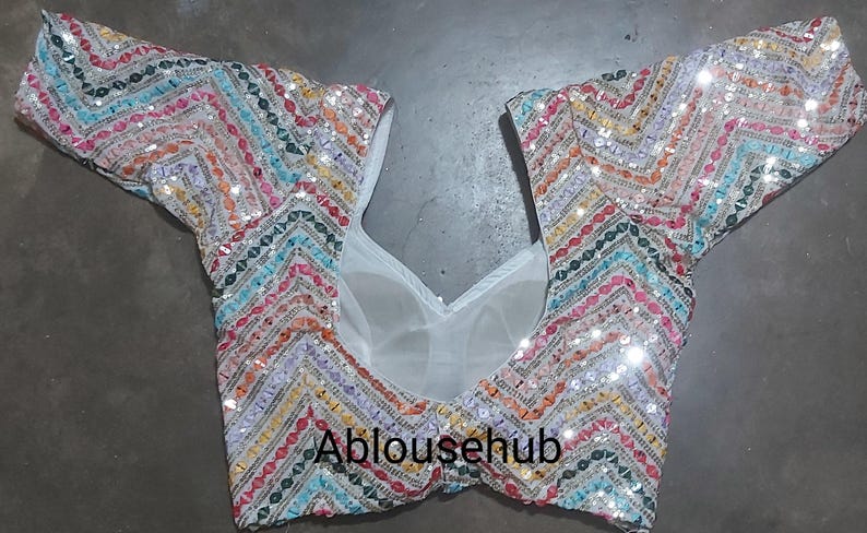 Puede incluir: Una blusa blanca con lentejuelas con un patr&oacute;n de chevron colorido. La blusa tiene un escote redondo y mangas cortas. El texto "Ablousehub" est&aacute; impreso en la parte delantera de la blusa.