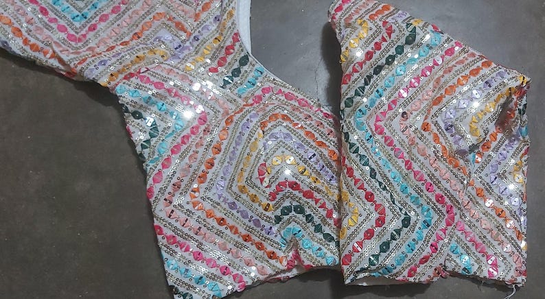 Puede incluir: Un top de tela blanca con un patr&oacute;n geom&eacute;trico colorido hecho de lentejuelas. El top tiene un escote en V y es sin mangas.