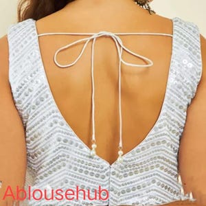 Puede incluir: Una blusa blanca sin mangas con un escote en V profundo y un cierre de lazo en la espalda. La blusa est&aacute; decorada con lentejuelas plateadas en un patr&oacute;n de chevron.