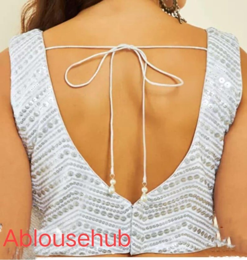Puede incluir: Una blusa blanca sin mangas con un escote en V profundo y un cierre de lazo en la espalda. La blusa est&aacute; decorada con lentejuelas plateadas en un patr&oacute;n de chevron.