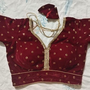 May include: A maroon velvet blouse with gold sequin accents. The blouse features a V-neckline and short sleeves, with gold trim along the edges and scattered gold sequins. A matching maroon headpiece is included.