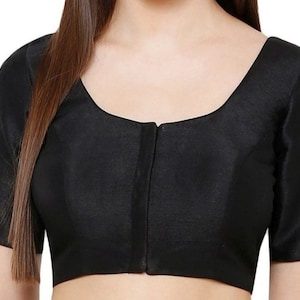 K&ouml;nnte beinhalten: Eine schwarze Bluse mit kurzen &Auml;rmeln und einem quadratischen Ausschnitt. Der Stoff hat einen subtilen Glanz, und eine vertikale Naht verl&auml;uft durch die Mitte der Vorderseite. Die Bluse ist einfarbig und scheint aus einem glatten Material zu sein.