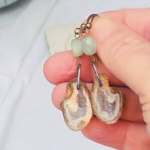 May include: Handmade earrings with natural stone slices and geode centres. The earrings are light brown and white, with small, light blue beads. They hang from silver-coloured wire. The earrings are a unique piece of jewellery.