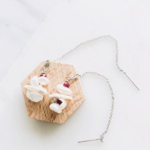 May include: A pair of silver dangle earrings with white pearl and red gemstone accents. The earrings are displayed on a wooden hexagonal stand.