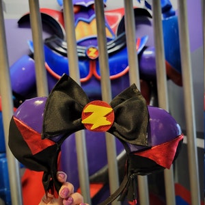 May include: Purple and black Minnie Mouse ears with a yellow lightning bolt design. The ears are held in front of a purple and blue figure in a cage.