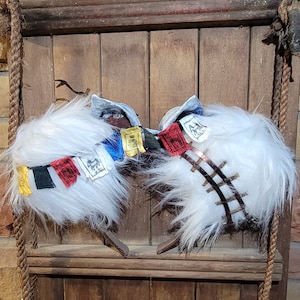 K&ouml;nnte beinhalten: Zwei wei&szlig;e, flauschige Schneeschuhe mit bunten Gebetsfahnen daran. Die Schneeschuhe h&auml;ngen an einer Holzwand mit einem Seil.