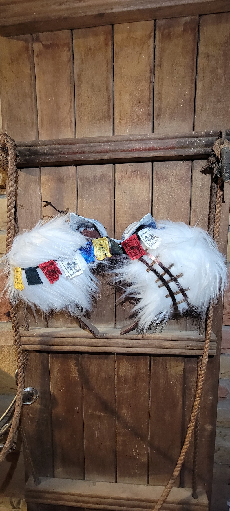 K&ouml;nnte beinhalten: Zwei wei&szlig;e, flauschige Schneeschuhe mit bunten Gebetsfahnen daran. Die Schneeschuhe h&auml;ngen an einer Holzwand mit einem Seil.