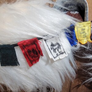 K&ouml;nnte beinhalten: Wei&szlig;es, flauschiges Garn mit einer Schnur aus bunten Stoffgebetfahnen. Die Fahnen sind gr&uuml;n, rot, wei&szlig;, blau und gelb. Die Fahnen haben verschiedene Symbole und Designs.