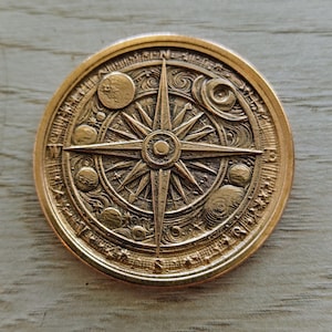 Può includere: Una moneta dettagliata color bronzo con bussola e un motivo a stella al centro. La moneta presenta elementi celesti, tra cui pianeti e stelle, ed è circondata da testo. La bussola indica i punti cardinali.