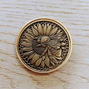 Op de afbeelding: Een ronde, antiek goudkleurige munt met een gedetailleerd reliëf van een bij op een zonnebloem. De bij is gepositioneerd over het midden van de zonnebloem, met ingewikkelde vleugel- en lichaamsdetails. De rand van de munt heeft een verhoogde rand.