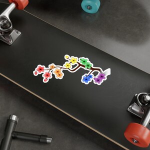 Pu&ograve; includere: Un ramo di ciliegio in fiore arcobaleno con uno stelo marrone e foglie verdi. I fiori sono disposti in fila, con i colori che passano dal rosso all'arancione, al giallo, al verde, al blu, all'indaco e al viola.